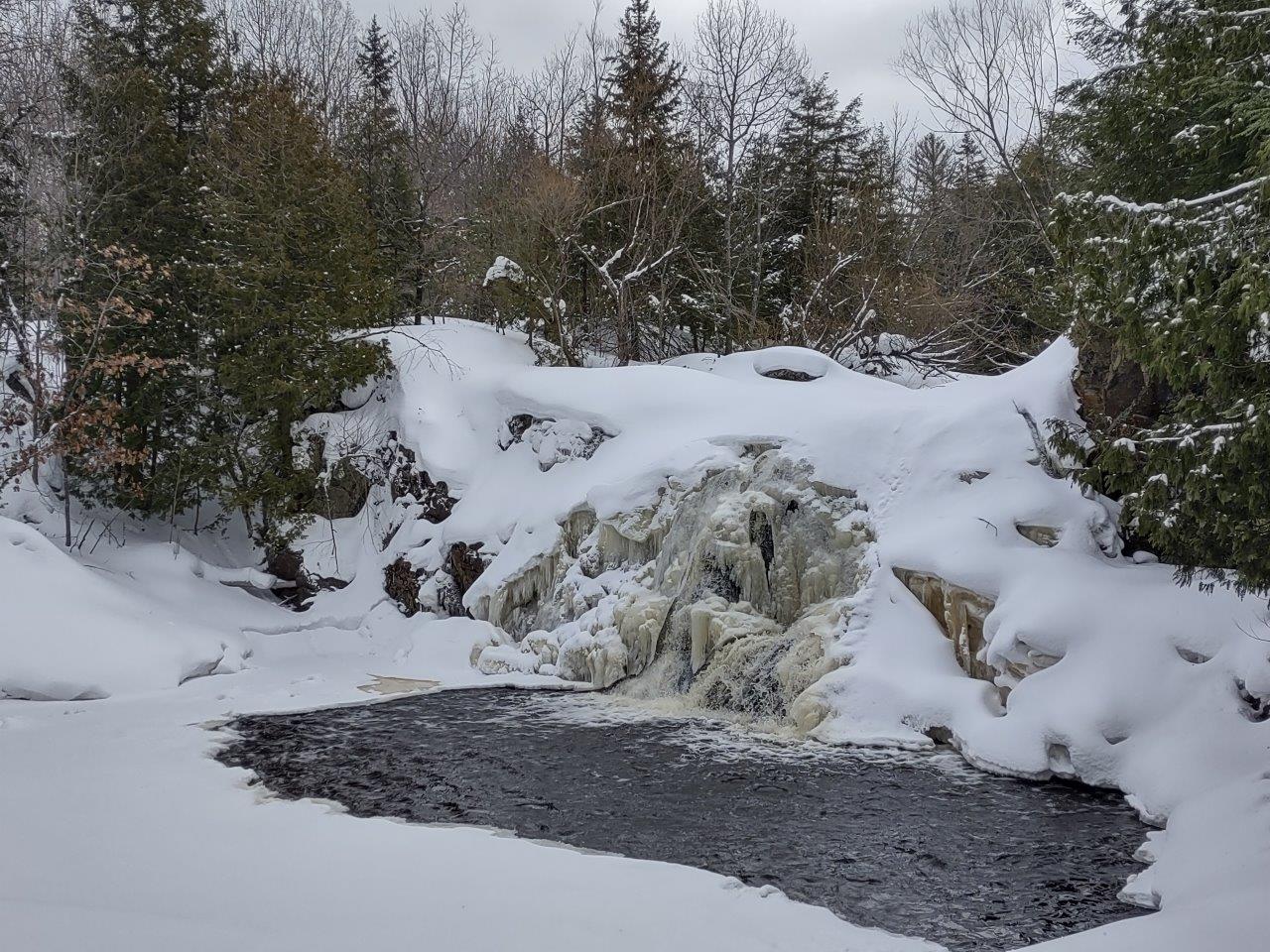 Ice covers much of the waterfall face, with water flowing under the ice. March 2022